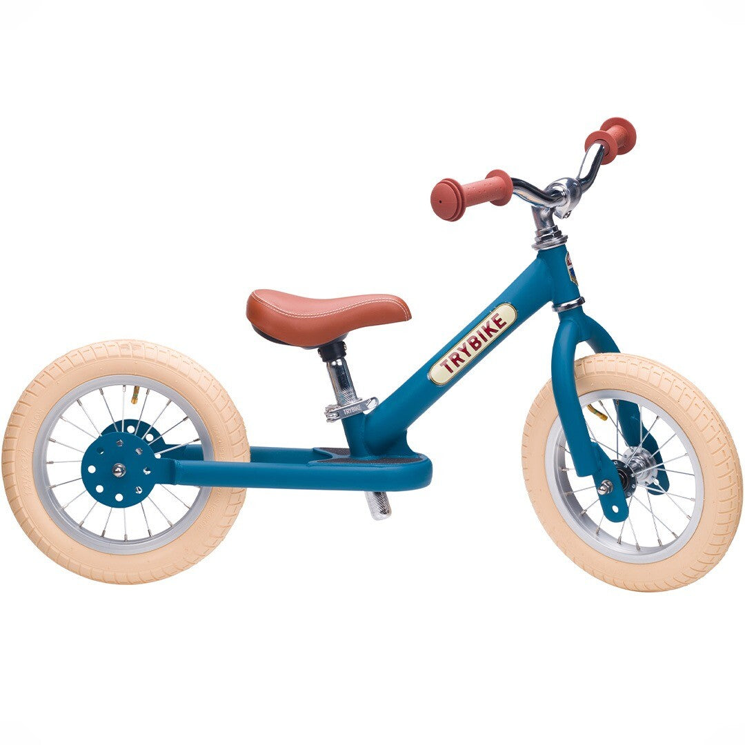 Trybike stalen loopfiets in vintage blauw met bruin zadel en crème banden, schuin van voren gefotografeerd.