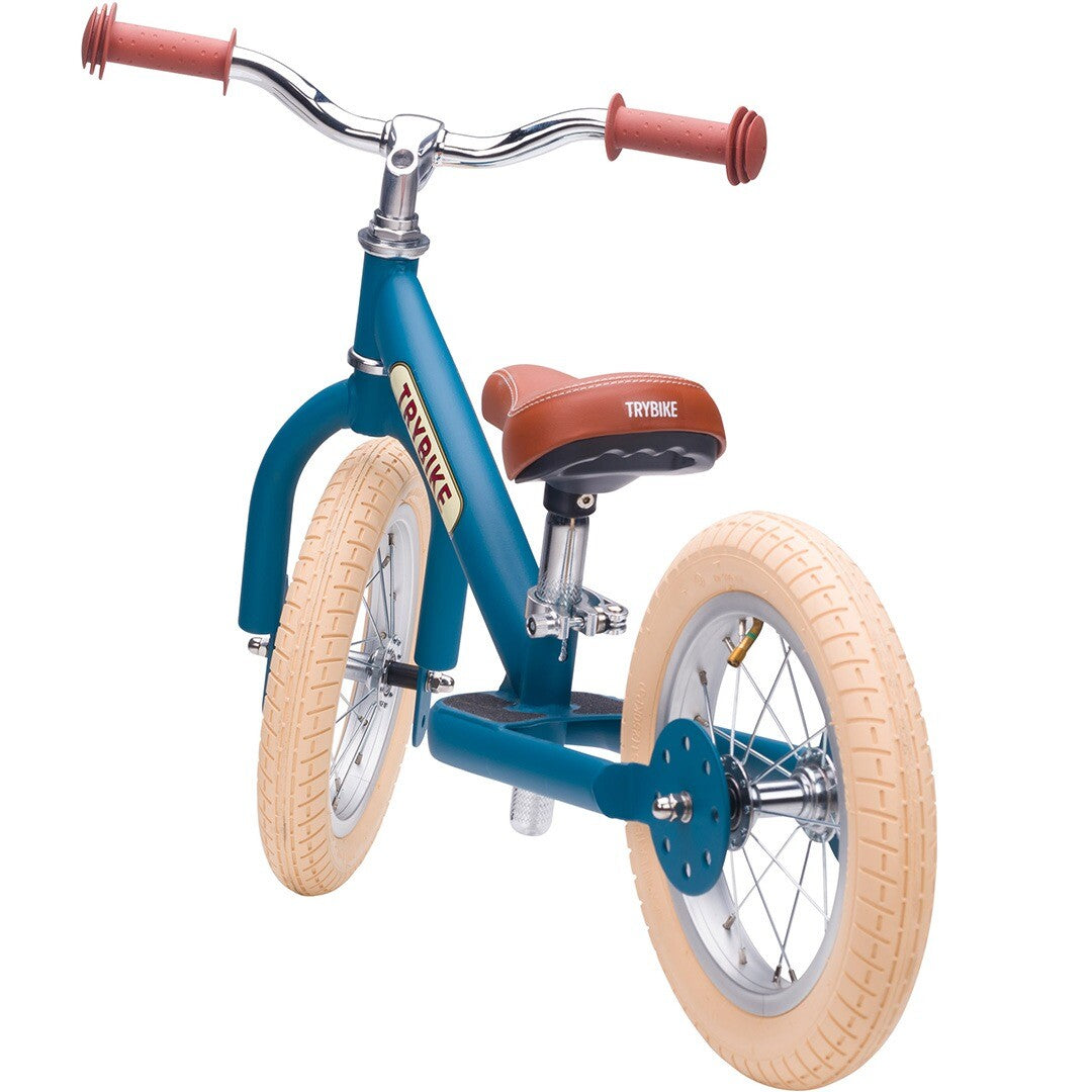 Trybike stalen loopfiets in vintage blauw met bruin zadel en crème banden, schuin van achteren gefotografeerd.