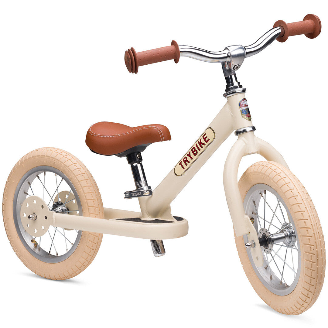 Trybike loopfiets in vintage cream met bruin zadel en handvatten, schuin van voren gefotografeerd.