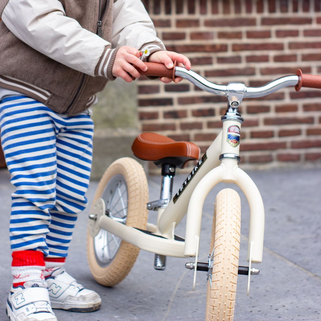 Een kind met gestreepte broek houdt de Trybike loopfiets vast, staand op een stoep voor een bakstenen muur.