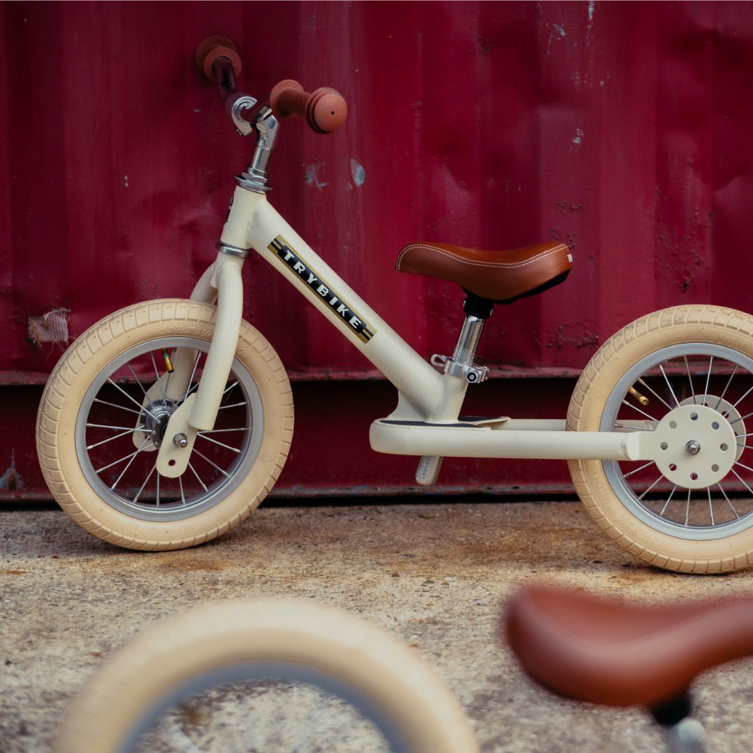 Trybike loopfiets in vintage cream, van de zijkant gezien, staand op beton voor een rode containerwand.