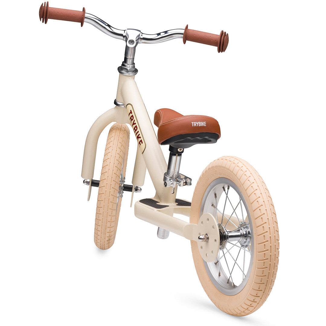 Trybike loopfiets in vintage cream met bruin zadel en handvatten, schuin van voren en laag gefotografeerd.