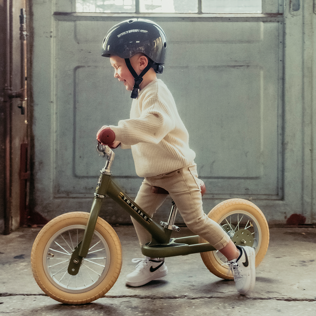 Een kind met helm rijdt lachend op een vintage groene Trybike stalen loopfiets.