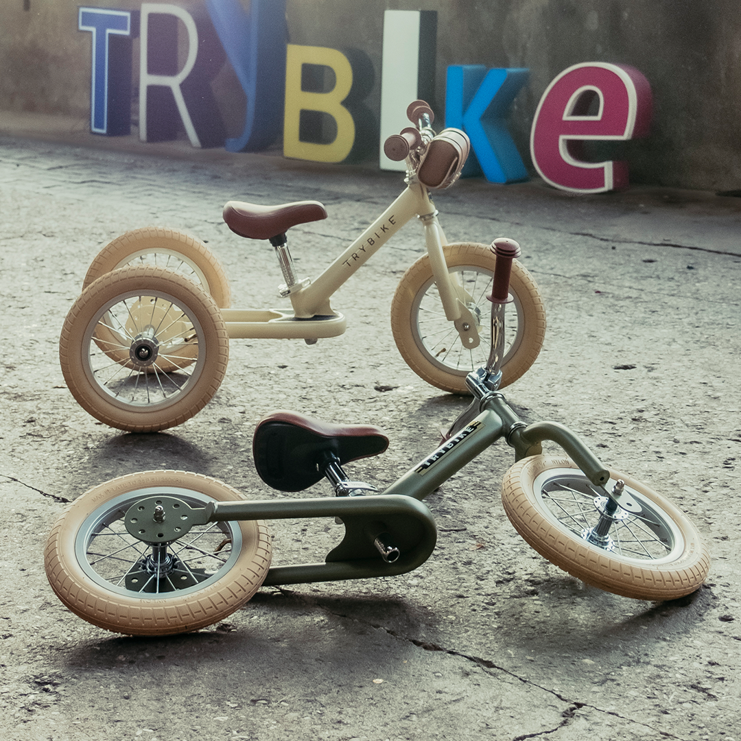 Twee Trybike loopfietsen, een crème driewieler en een vintage groene tweewieler, staan op een betonnen vloer.