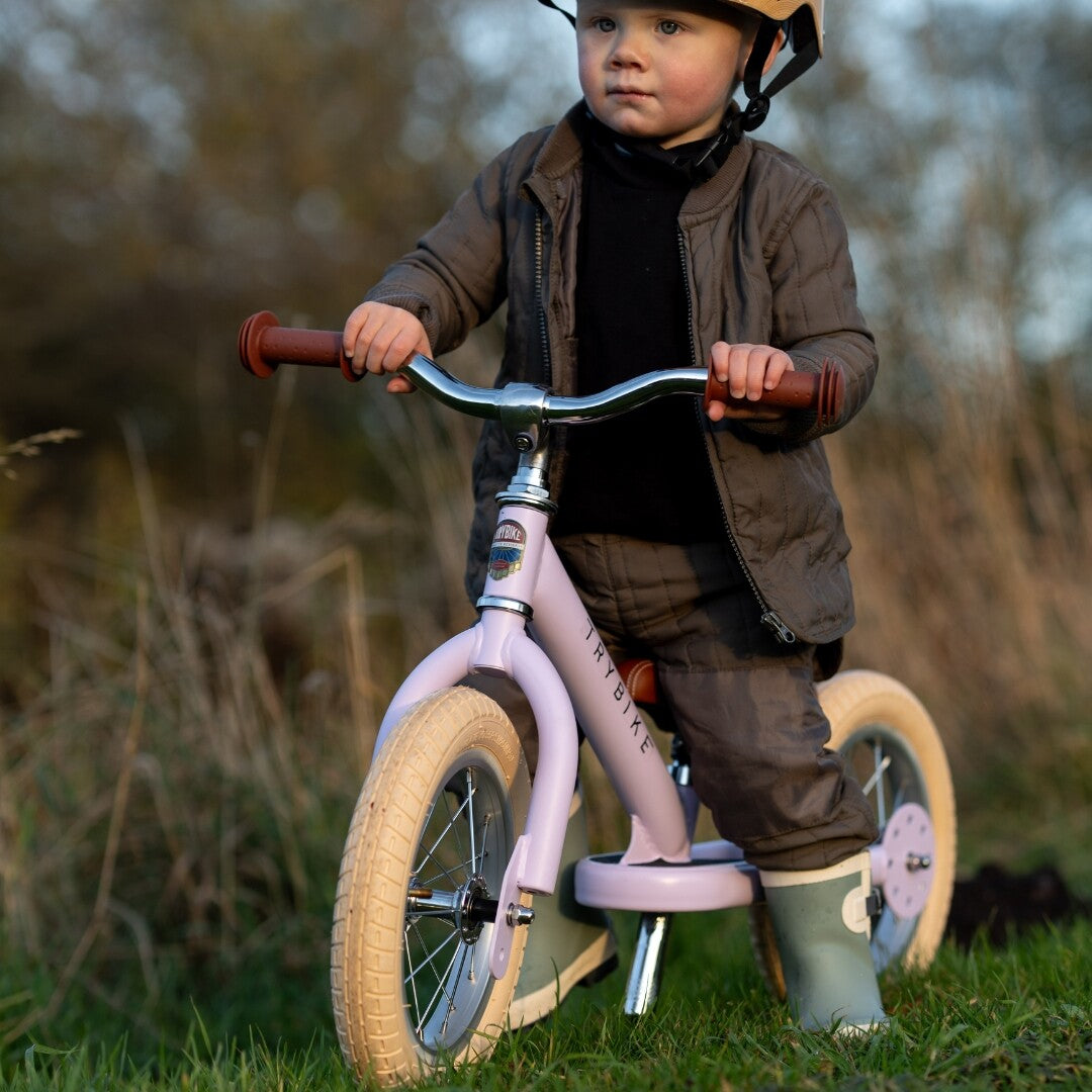 Een kind met helm staat buiten in het gras naast de lila Trybike loopfiets.
