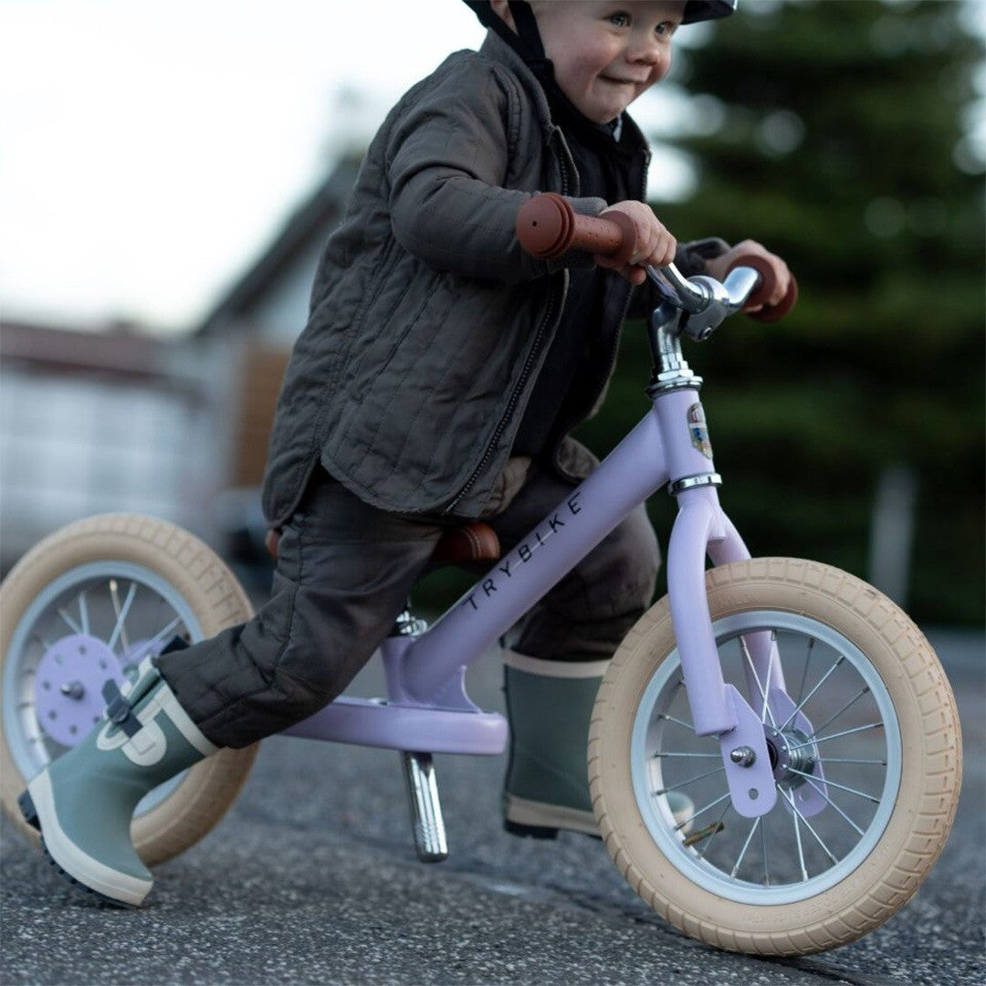 Een lachend kind met helm rijdt op de lila Trybike loopfiets over een pad.