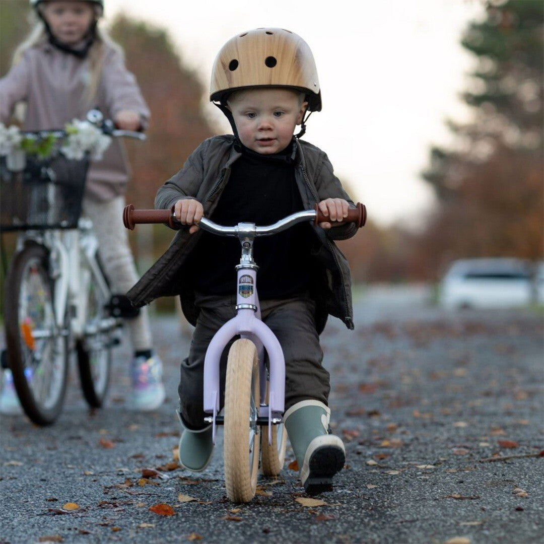 Een kind met helm rijdt op de lila Trybike loopfiets op een pad, met een ander kind op de achtergrond.