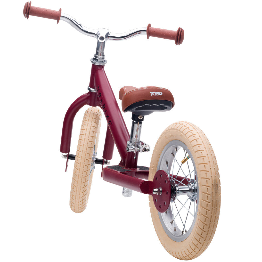 Trybike stalen loopfiets in vintage rood met bruin zadel en crèmekleurige banden, schuin van achteren gefotografeerd.