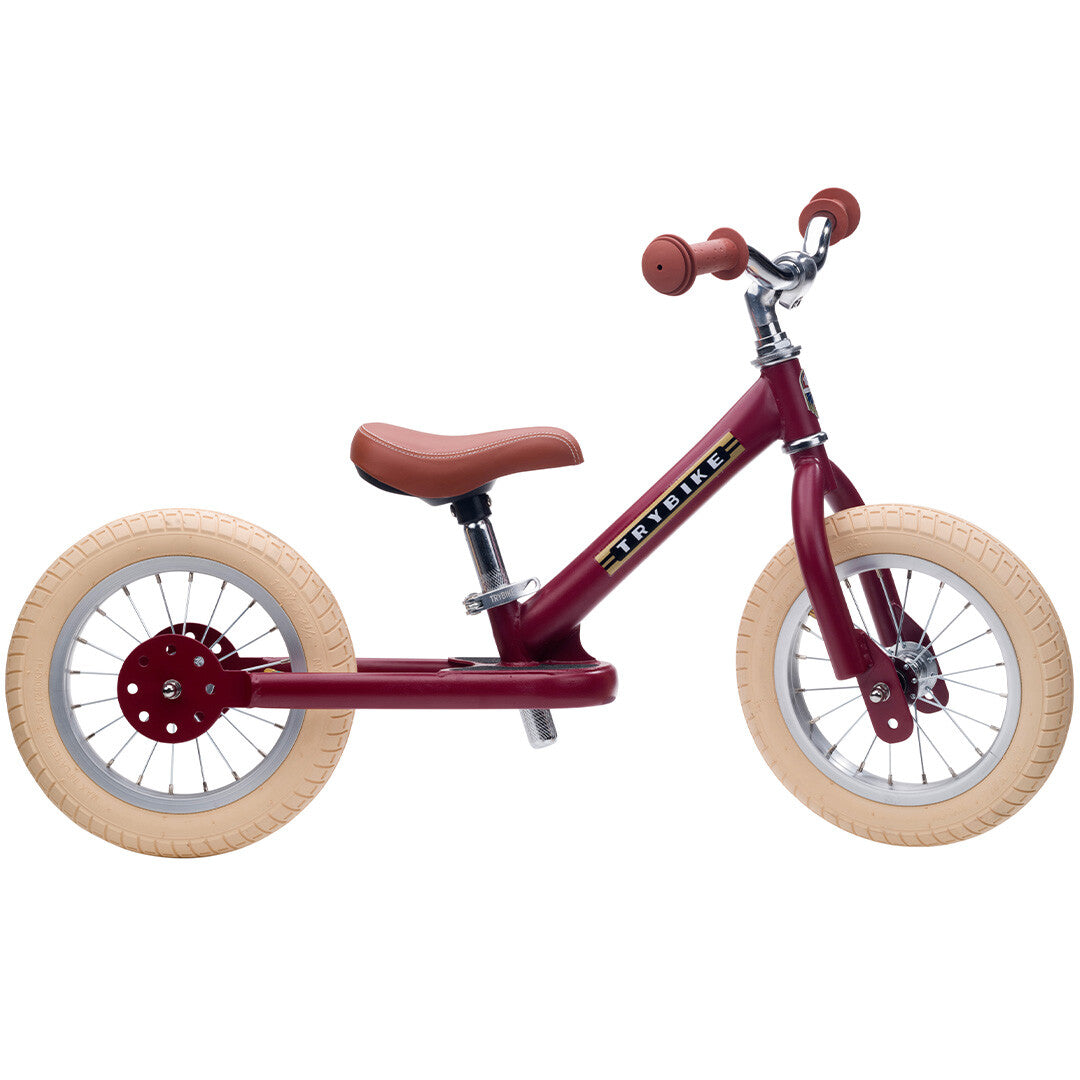 Trybike stalen loopfiets in vintage rood met bruin zadel en crèmekleurige banden, van de zijkant gefotografeerd.