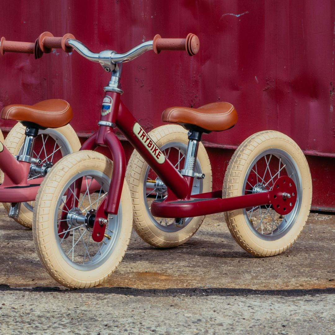 Twee Trybike stalen loopfietsen, een vintage rode en een roze, staan naast elkaar voor een rode muur.