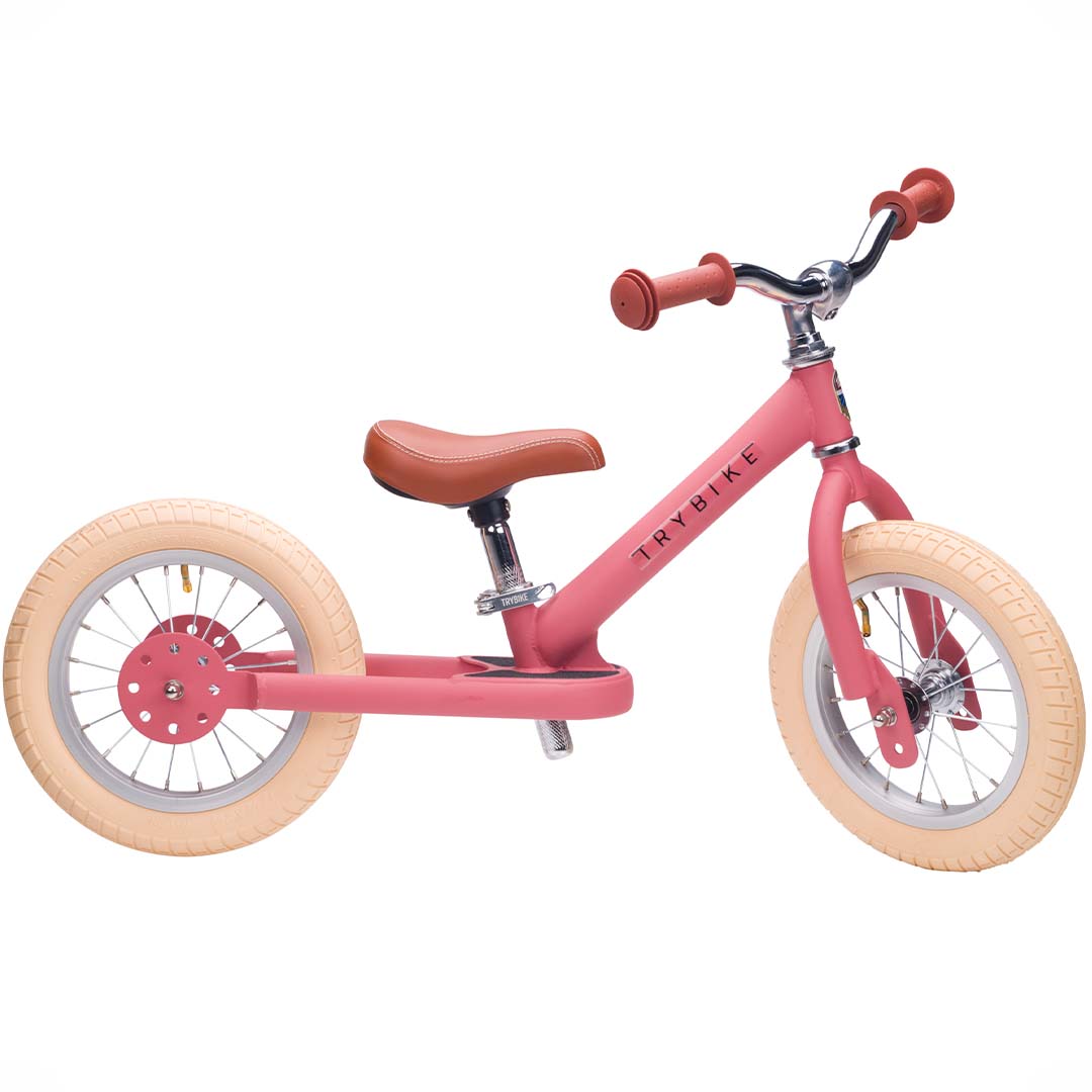 Trybike stalen loopfiets in vintage roze met bruin zadel en lichte banden, schuin van opzij gefotografeerd.