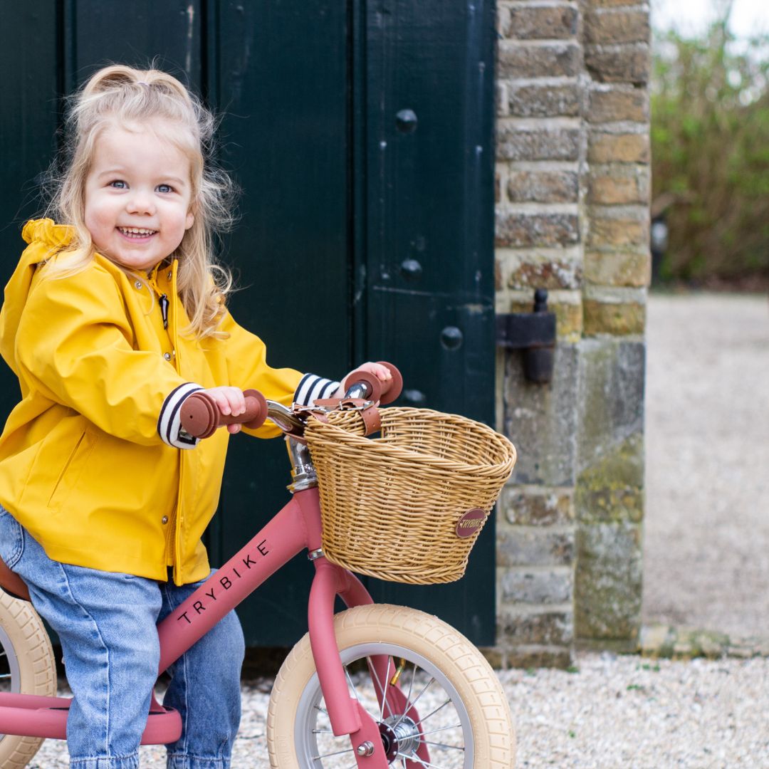 Lachend kind in gele regenjas op een Trybike vintage roze loopfiets met rieten mandje voor een oude muur.