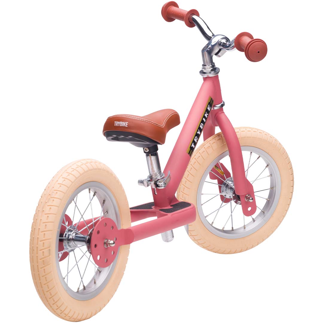 Trybike stalen loopfiets in vintage roze met bruin zadel en lichte banden, schuin van achteren gefotografeerd.