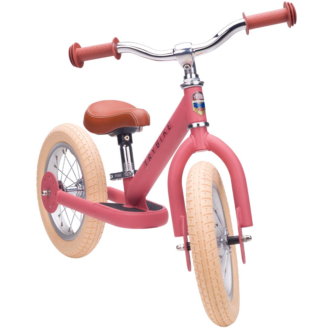 Trybike stalen loopfiets in vintage roze met bruin zadel en lichte banden, schuin van voren gefotografeerd.