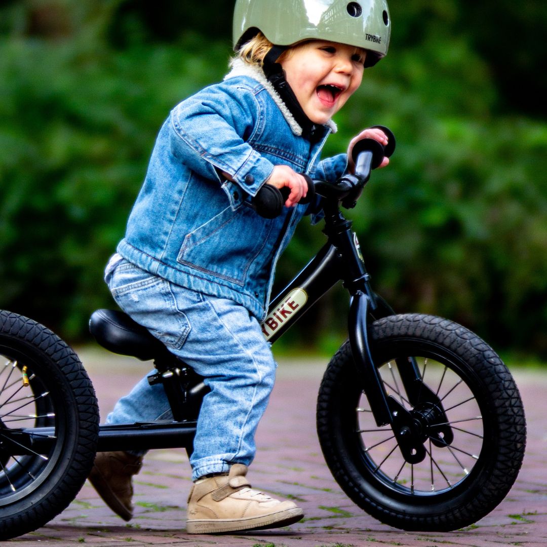 Blij kind met helm op een zwarte Trybike stalen loopfiets rijdend op een pad.
