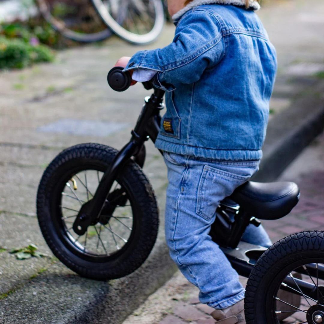 Kind van achteren gezien op een zwarte Trybike stalen loopfiets rijdend langs een stoeprand.