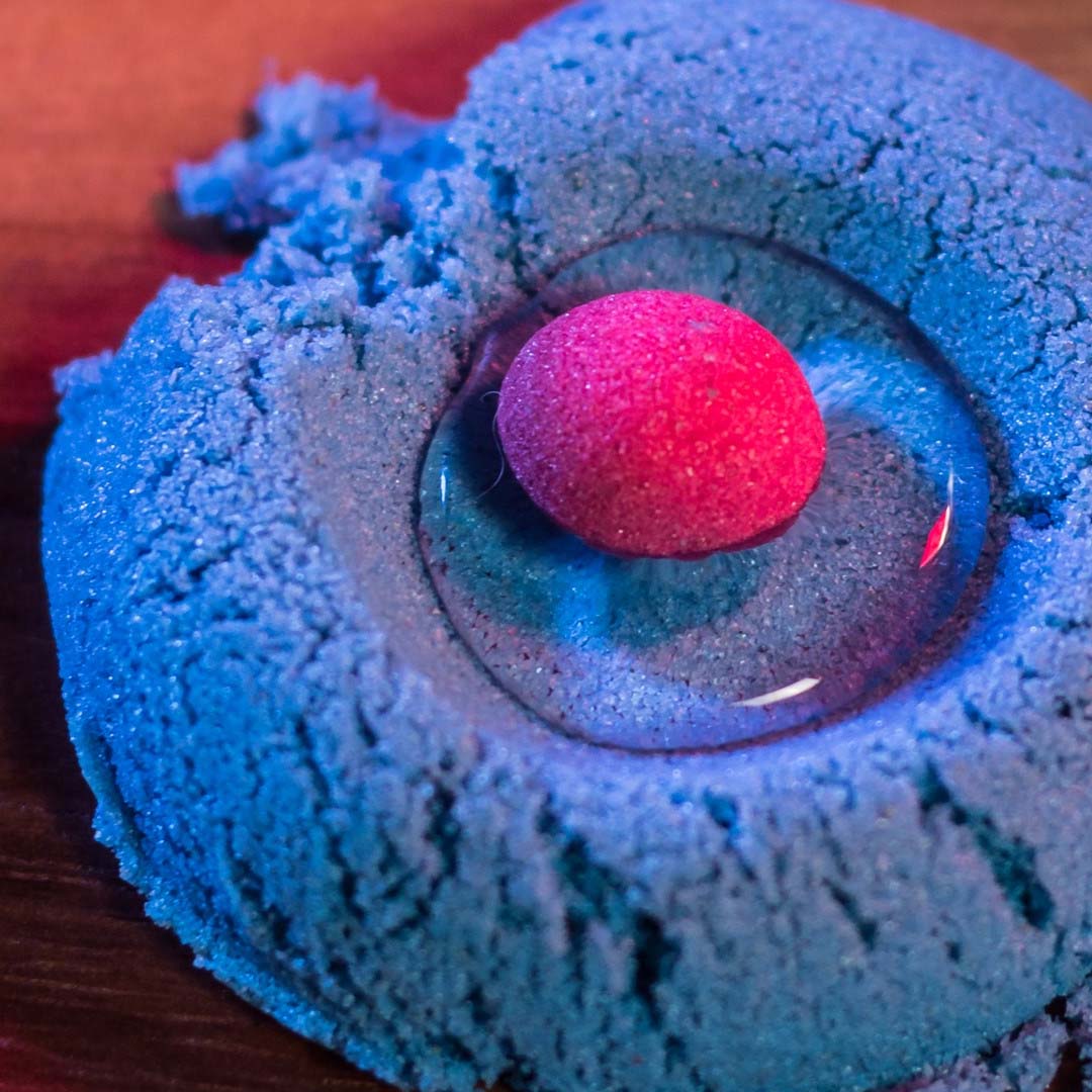 Detail van blauw kinetisch zand met een roze bal in een plasje heldere vloeistof.