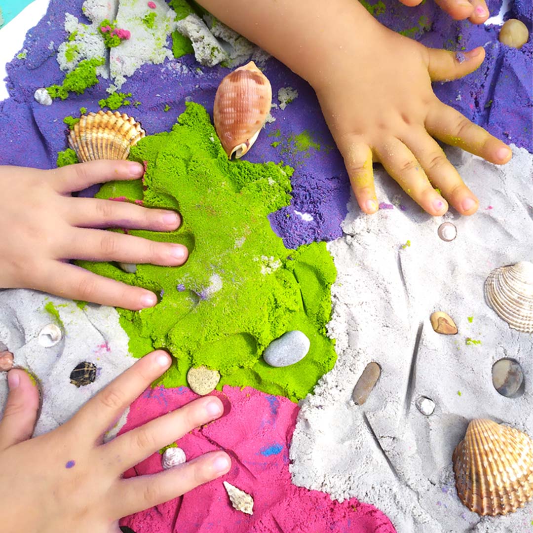 Twee kinderhanden spelen met Tuban kinetisch zand in verschillende kleuren, met schelpen en steentjes.