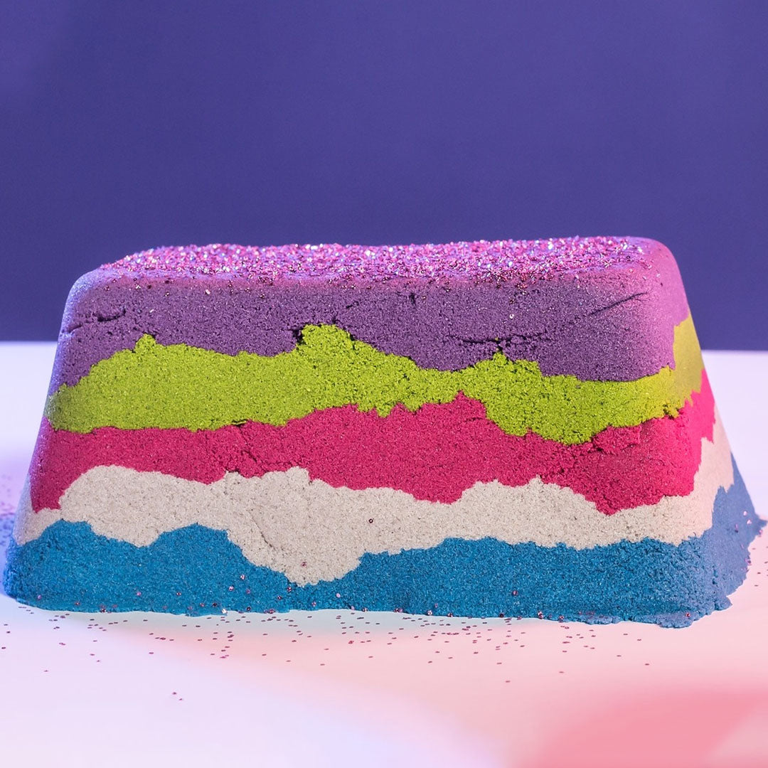 Een blok kinetisch zand met lagen in paars, groen, roze, wit en blauw, bestrooid met glitter.