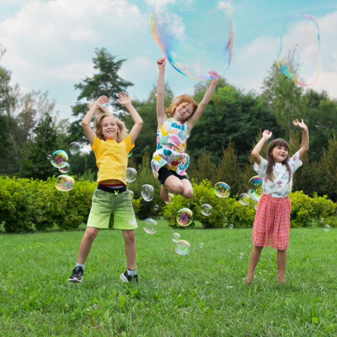 Drie kinderen springen vrolijk in een grasveld tussen grote en kleine bellen.