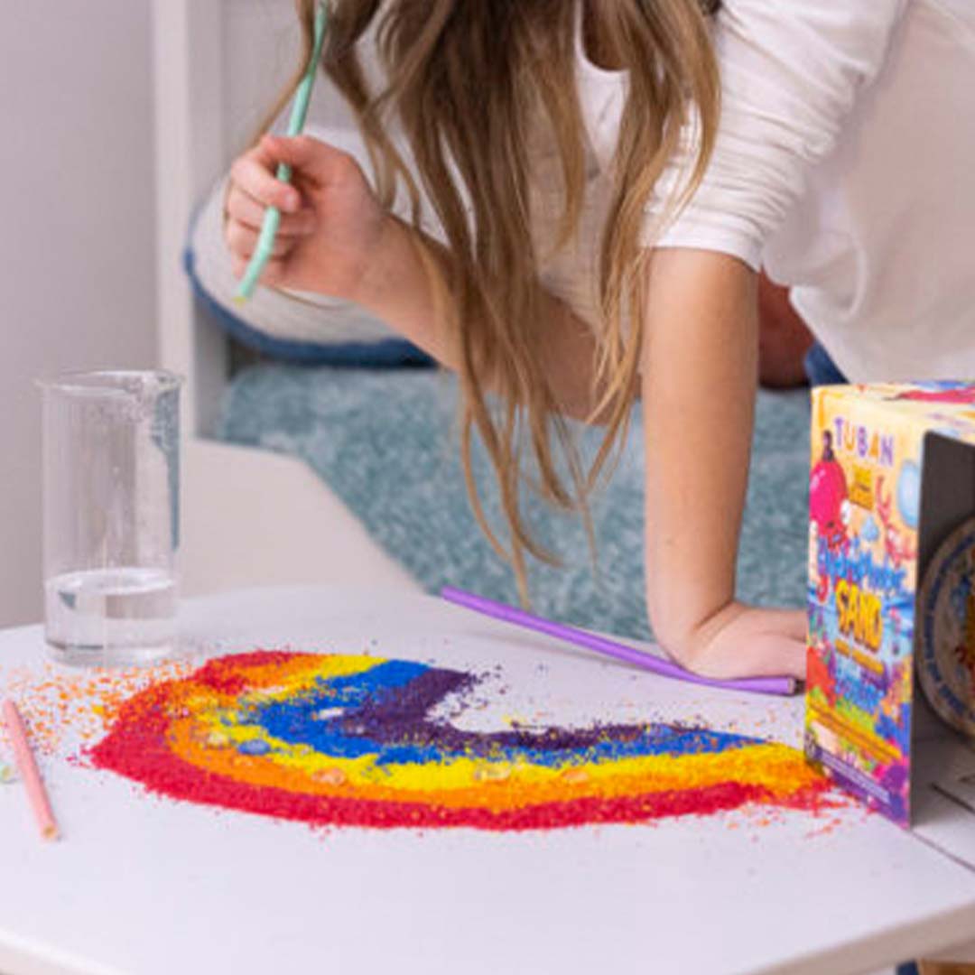 Een kind maakt een regenboogpatroon met kleurrijk hydrofoob zand op een tafel.