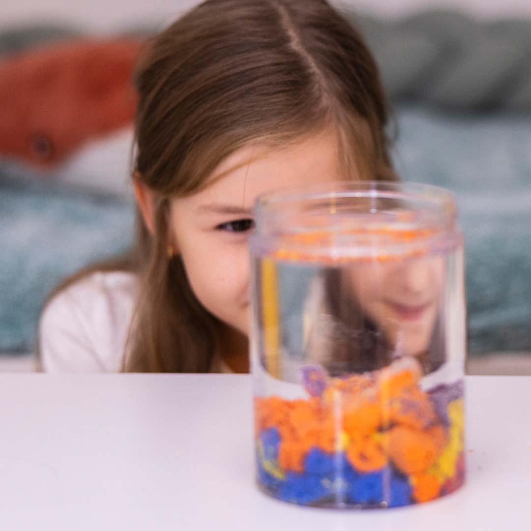 Een kind kijkt naar kleurrijk hydrofoob zand in een doorzichtige pot met water.