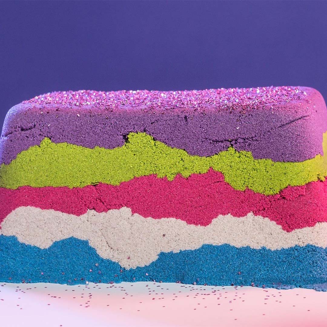 Een blok van gelaagd kinetisch zand in paars, groen, roze, grijs en blauw, bestrooid met glitters.