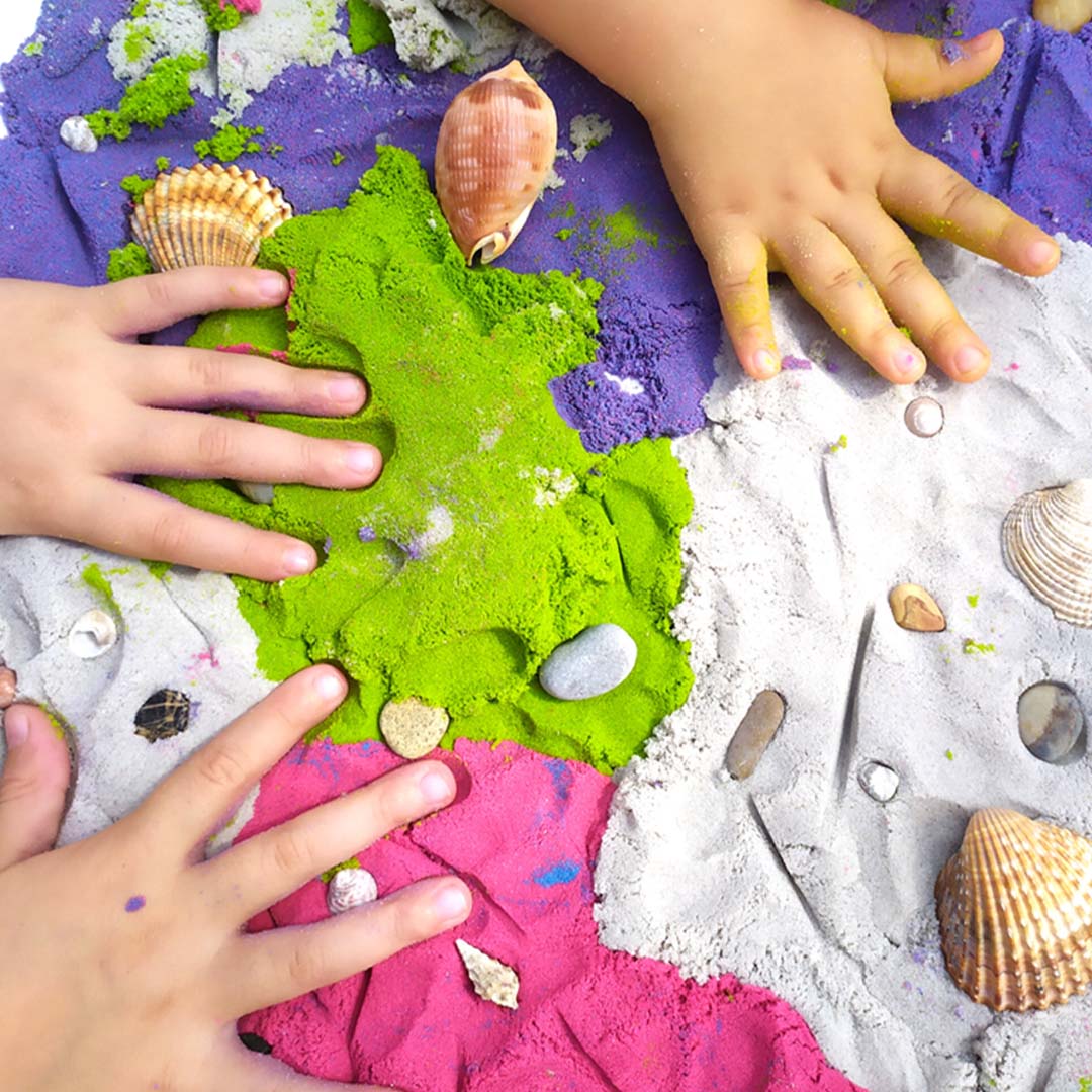 Kinderhanden spelen met groen, paars, roze en beige kinetisch zand en schelpen.