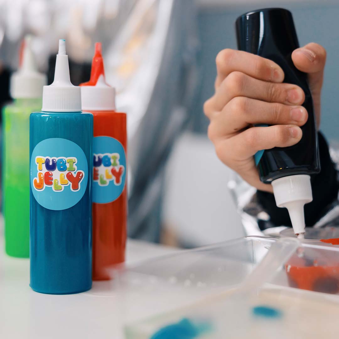 Een hand knijpt zwarte Tubi Jelly vloeistof uit een fles in een bakje, met andere kleuren Tubi Jelly flessen op de achtergrond.
