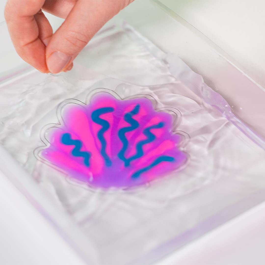 Een hand tilt een roze en blauwe schelpvormige Tubi Jelly figuur uit het water.