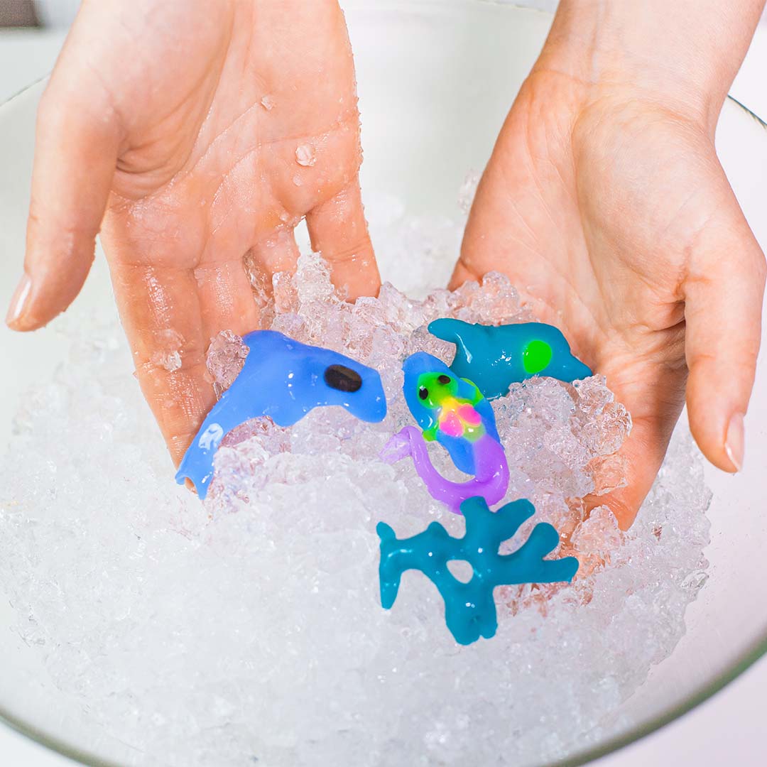 Twee handen houden verschillende kleurrijke Tubi Jelly onderwaterdieren boven een kom met ijsschilfers.