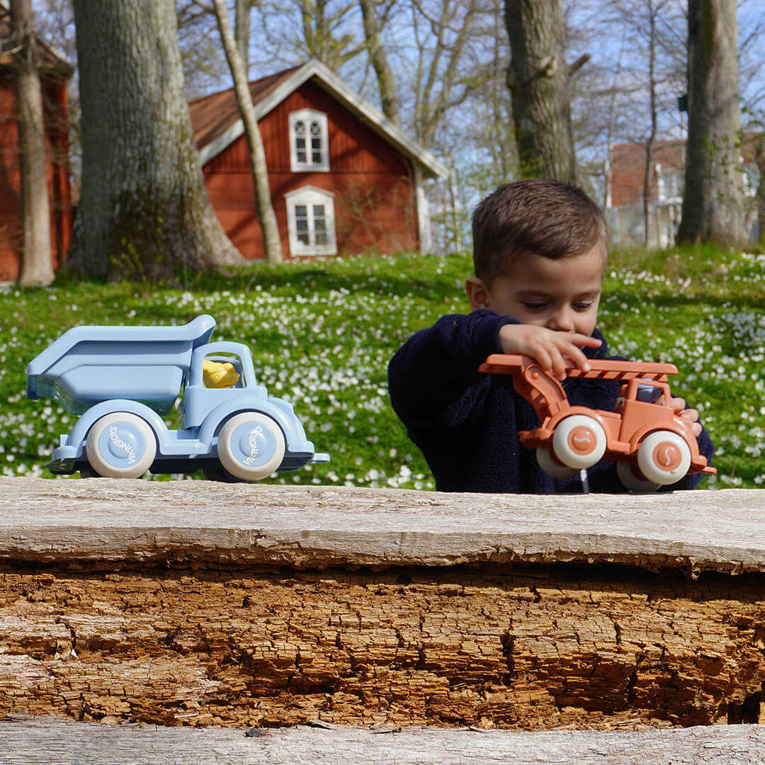 Een jongen speelt buiten met een blauwe Viking Toys kiepwagen en een oranje speelgoedvoertuig op een boomstam.