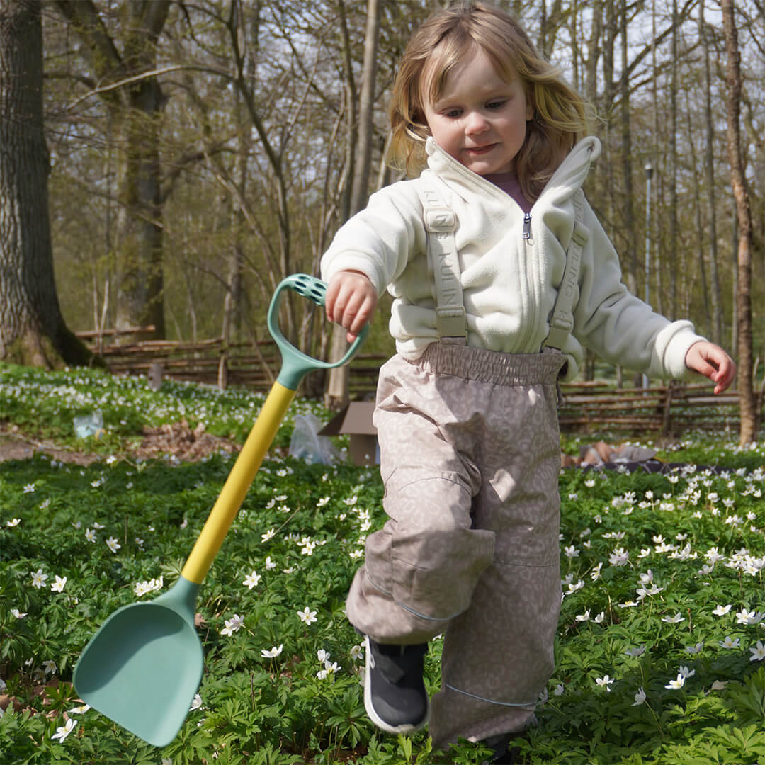 Een kind loopt met de Viking Toys re:line zandschep door een bos vol witte bloemen.