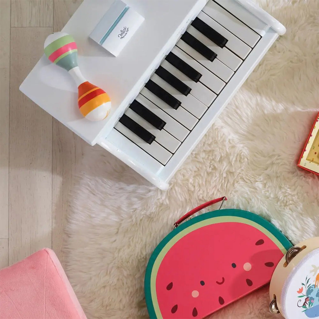 Een speelgoedset met een witte piano, een gestreepte maraca, een watermeloen tas en een tamboerijn op een witte vloerkleed.
