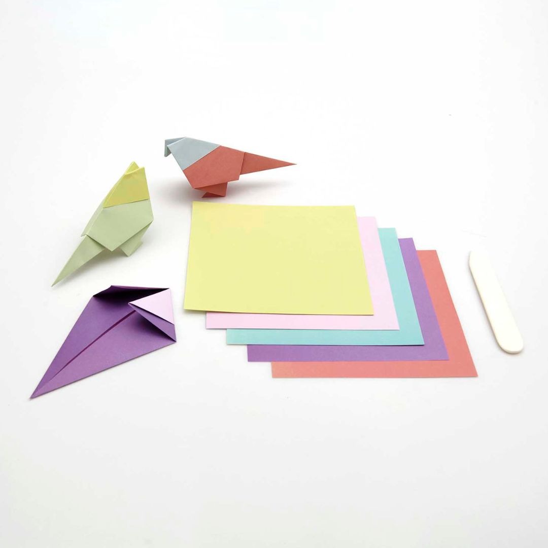 Diverse vellen kleurrijk origamipapier, deels gevouwen tot vogels, met een vouwbeen.