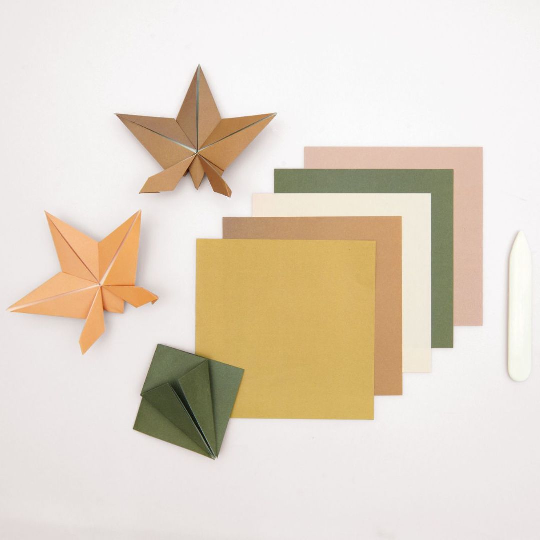 Vellen origamipapier in aardetinten met twee gevouwen sterren en een vouwbeen op een witte ondergrond.