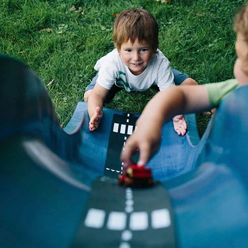 Een lachende jongen speelt met een rood speelgoedautootje op een zwart waytoplay rubberen wegdeel op een blauwe glijbaan.