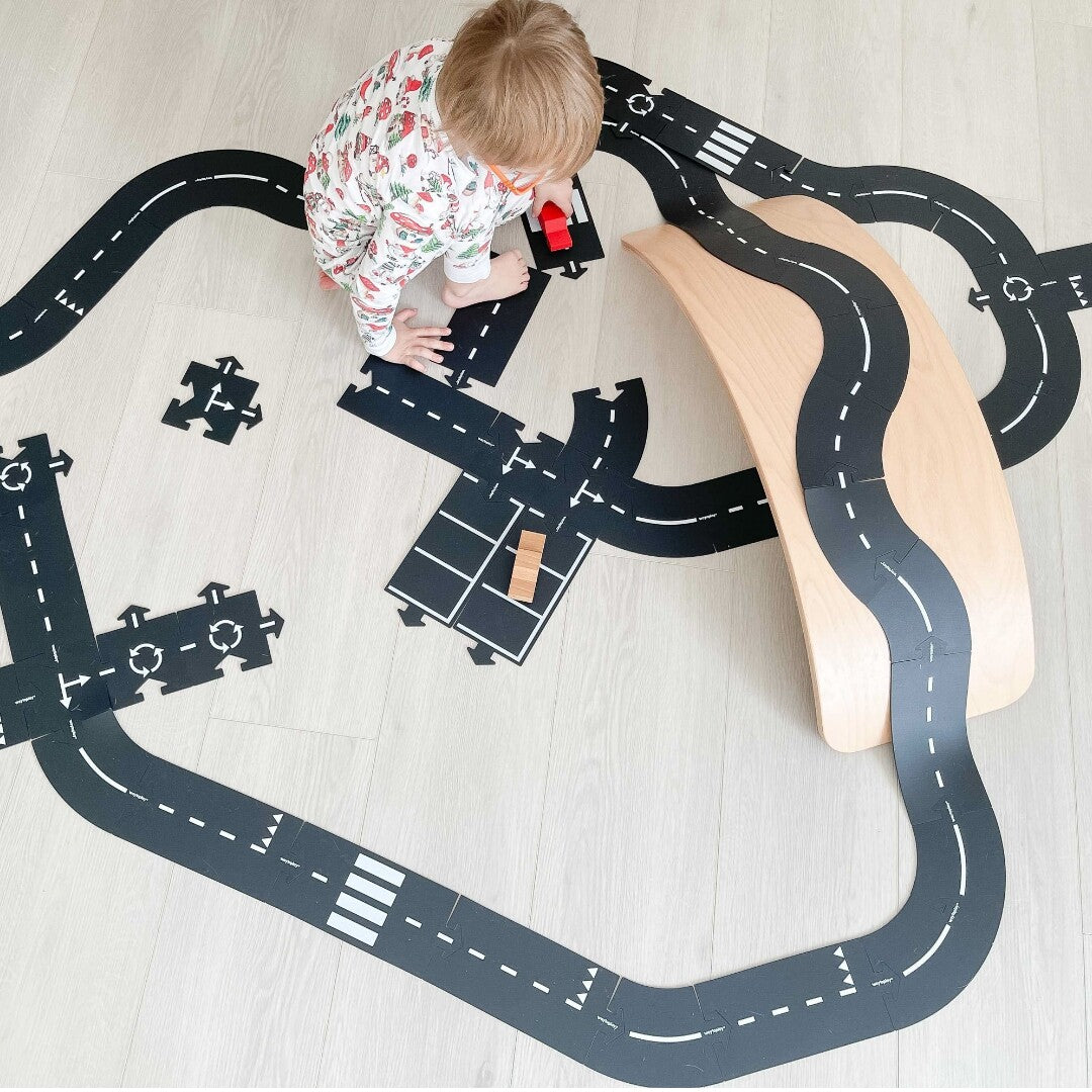Een kind speelt op de vloer met een grote waytoplay rubberen weg en een houten balansbord als brug.