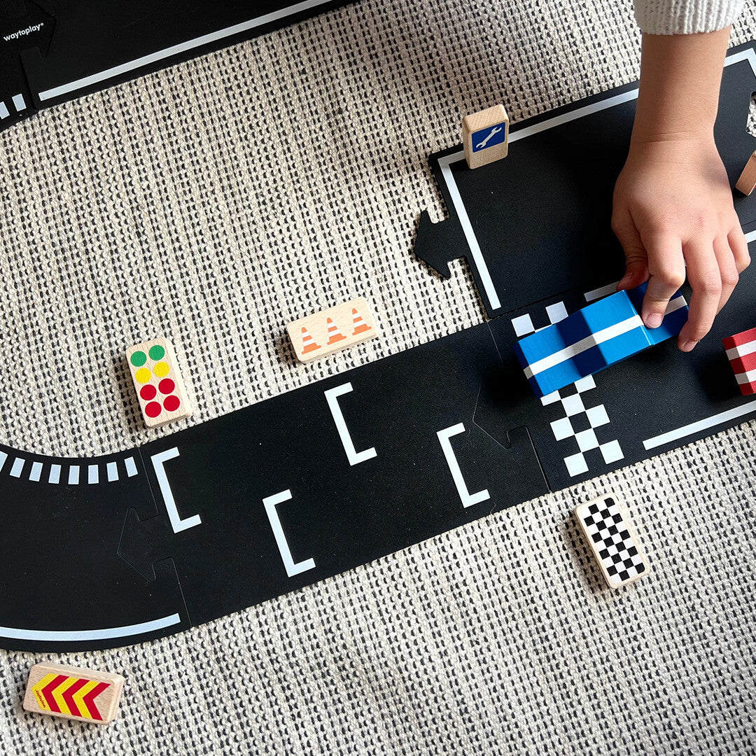 Een kinderhand beweegt een blauwe speelgoedauto over een zwarte racebaan met waytoplay houten raceborden.