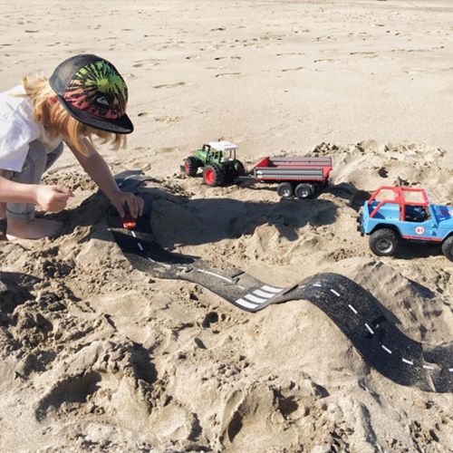 Een kind speelt met waytoplay rubberen wegdelen en speelgoedvoertuigen in het zand op het strand.