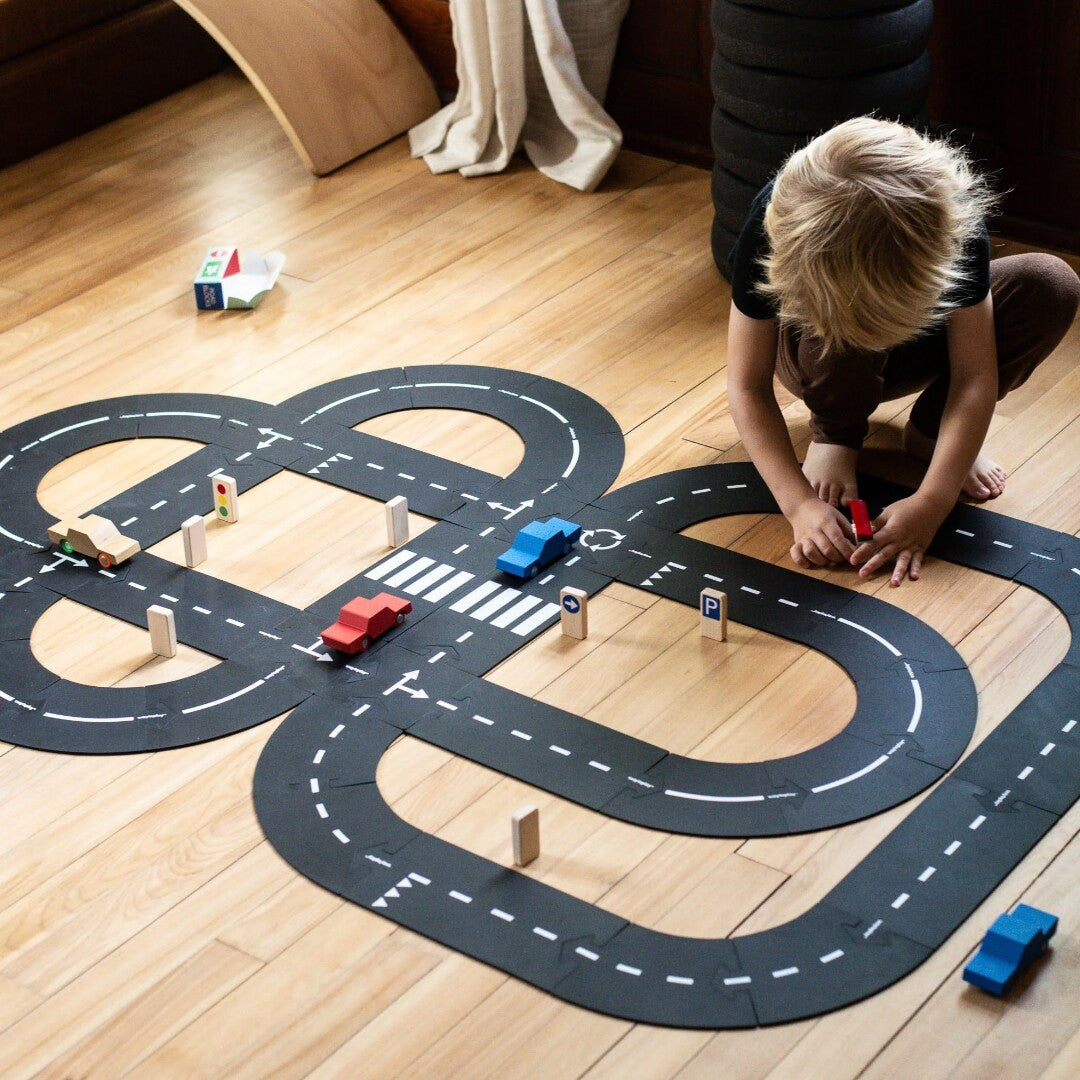 Een kind speelt op de grond met een grote waytoplay wegmat, houten auto's en verkeersborden.