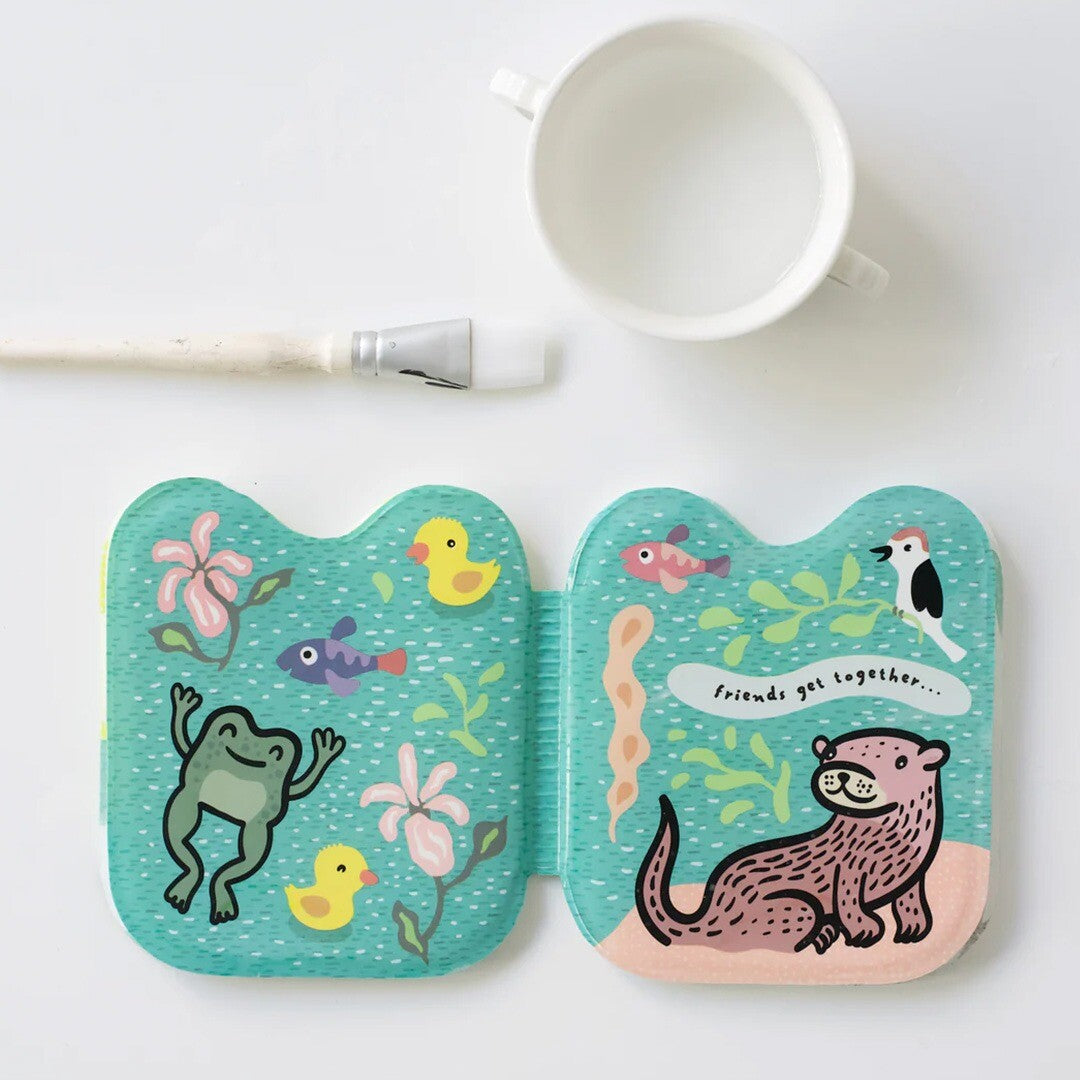Open badboekje met gekleurde illustraties van een kikker, eendjes, vissen en een otter, met een kwast en een beker ernaast.