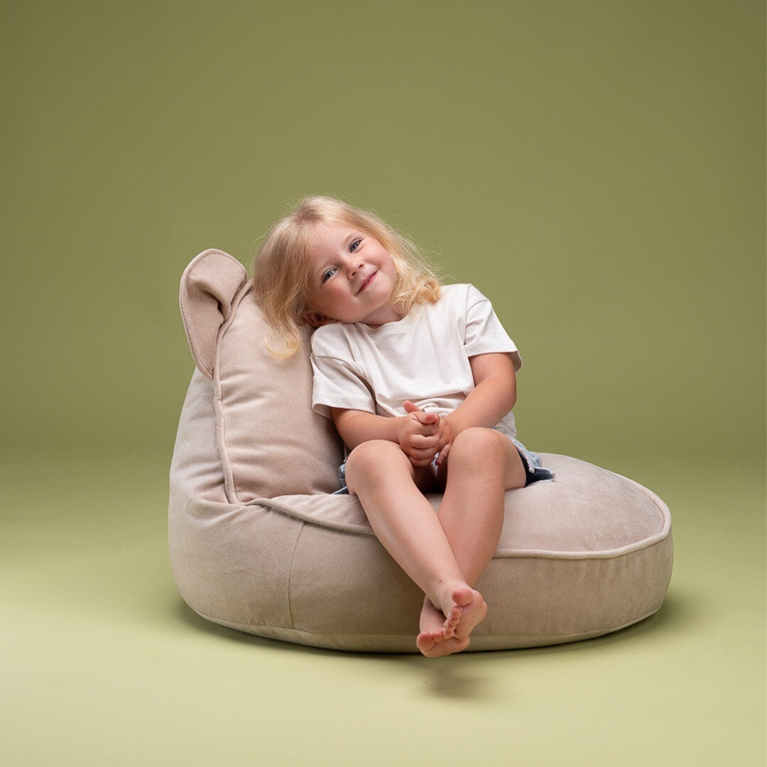 Een lachend blond meisje zit comfortabel in de wigiwama beige fluwelen zitzak met berenoren.