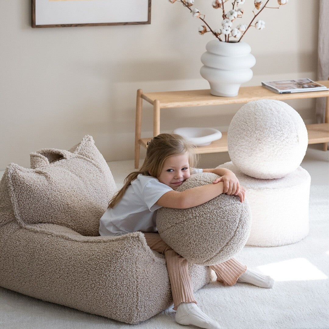 Een lachend meisje zit in de Wigiwama beige kinderzitzak stoel en knuffelt een rond kussen.