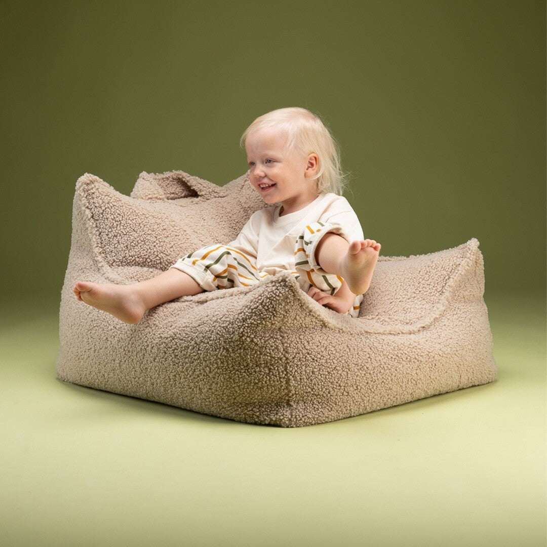 Een lachende peuter zit in de Wigiwama beige kinderzitzak stoel op een groene achtergrond.