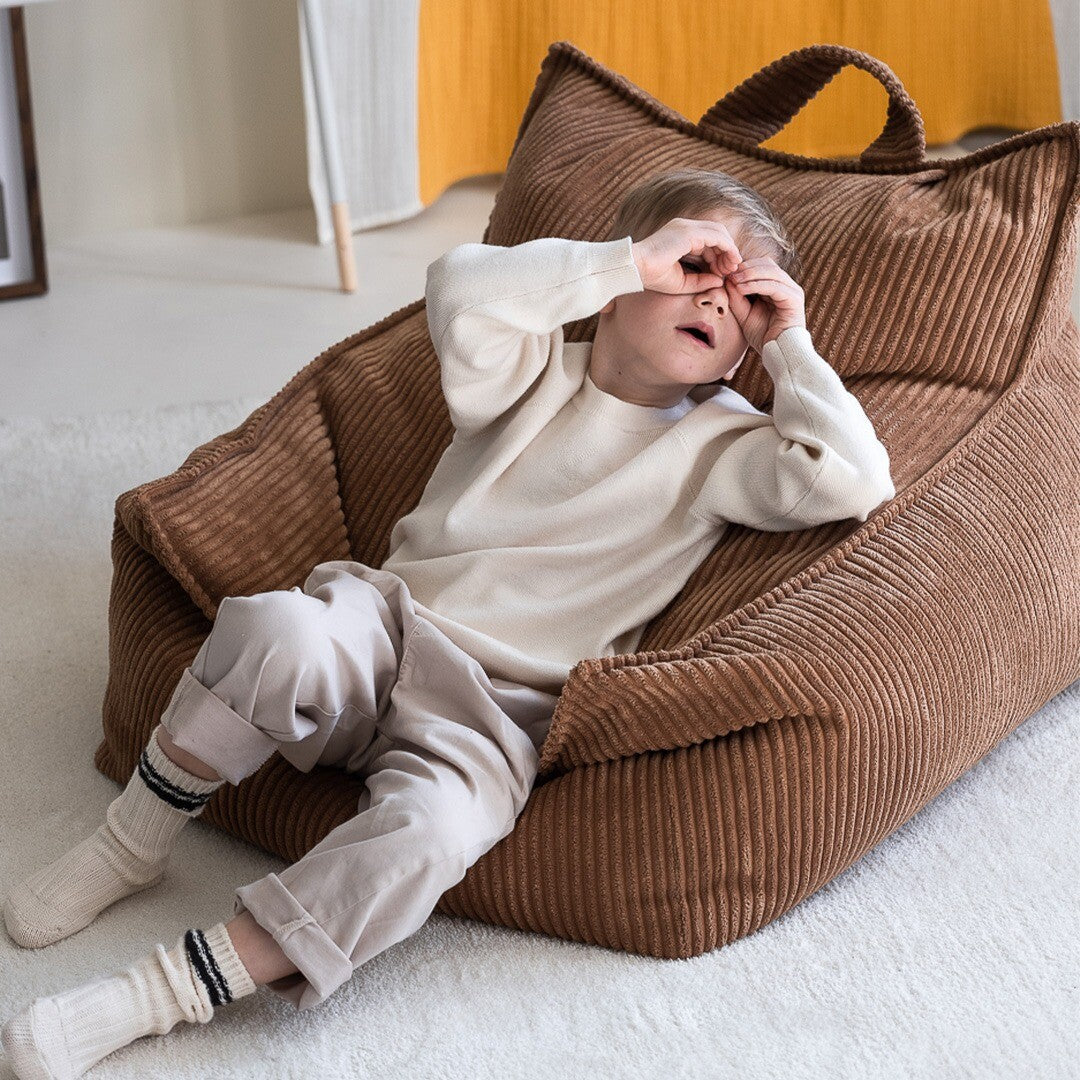 Een jongen zit in de wigiwama zitzak stoel van bruin corduroy en bedekt zijn ogen met zijn handen.