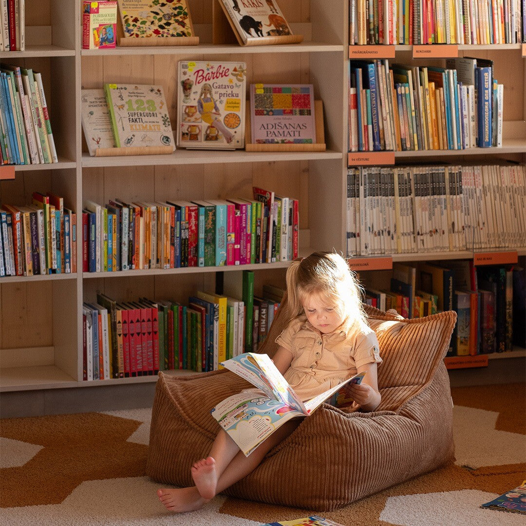 Een meisje leest een boek terwijl ze zit in de wigiwama zitzak stoel van bruin corduroy voor een boekenplank.