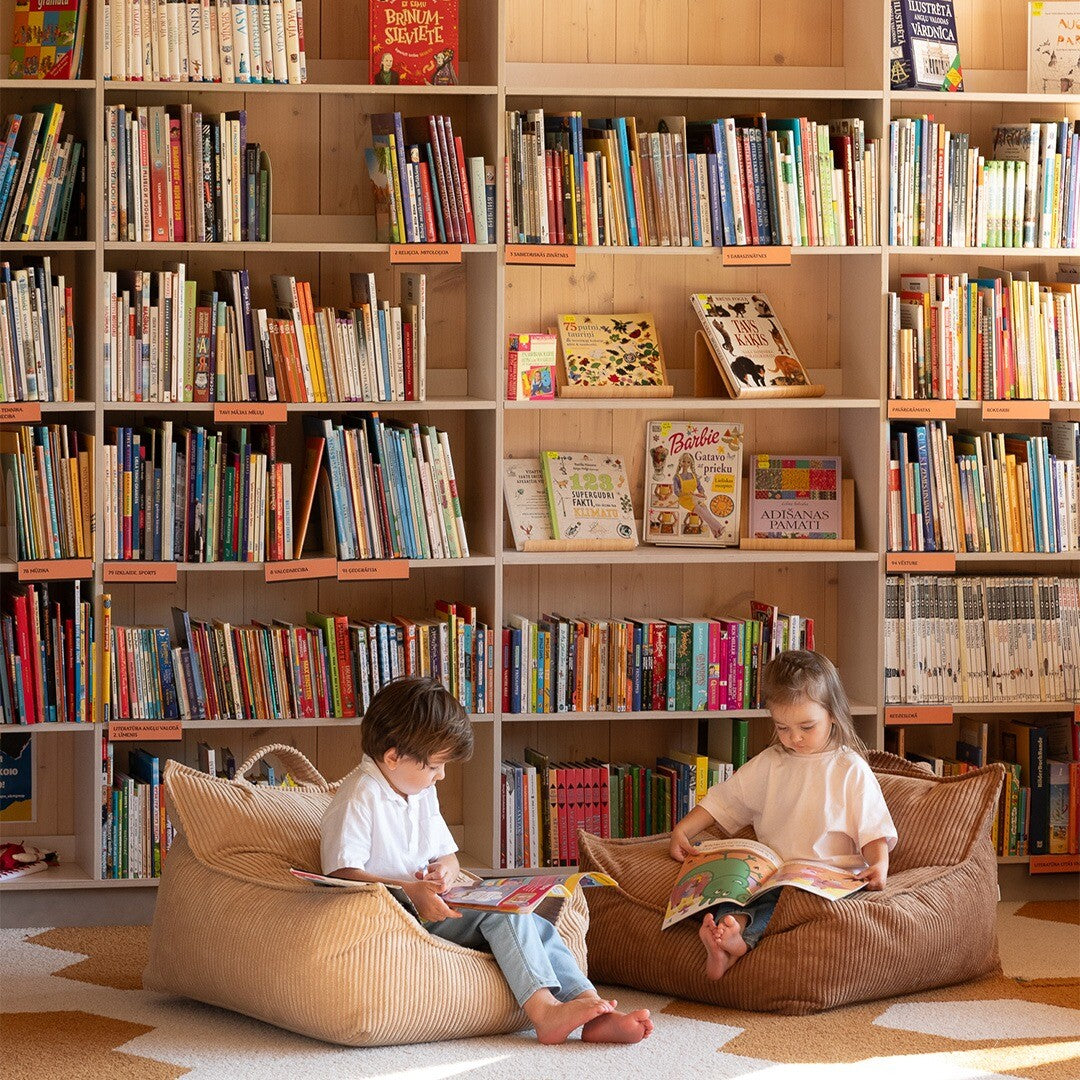 Twee kinderen lezen boeken in twee wigiwama zitzak stoelen van bruin corduroy voor een boekenplank.