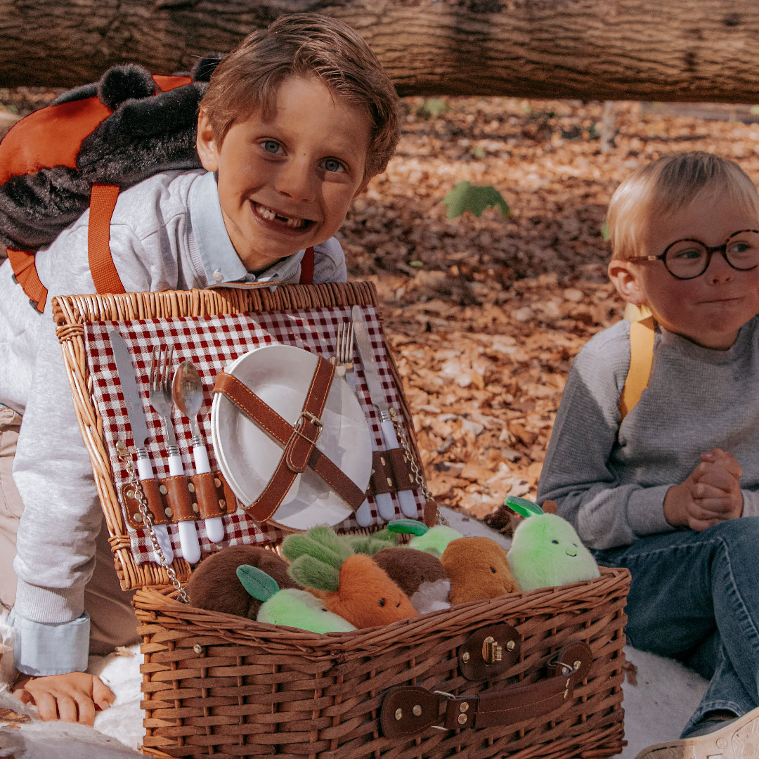 Twee kinderen zitten bij een open picknickmand met pluchen groente- en fruitknuffels in een herfstbos.
