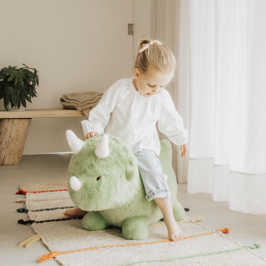 Een jong meisje stapt op het Wild & Soft groene pluche skippydier in de vorm van een dinosaurus, binnen op een vloerkleed.